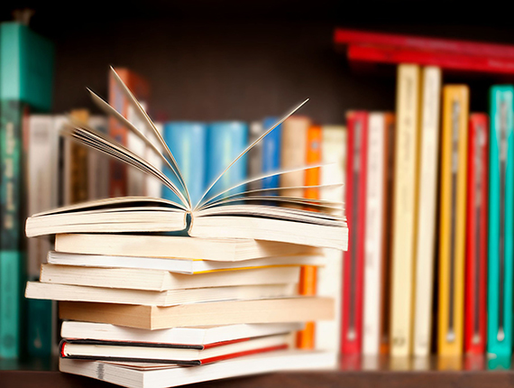 Stack of books on a wooden library shelf, one of them open on top, multicolored book spines background.